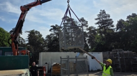 Ein Teilnehmer platziert eine Gitterbox präzise über zwei anderen, während ein Einweiser den Prozess mit einer Holzstange unterstützt.