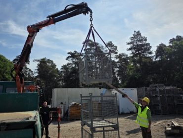 Ein Teilnehmer platziert eine Gitterbox präzise über zwei anderen, während ein Einweiser den Prozess mit einer Holzstange unterstützt.