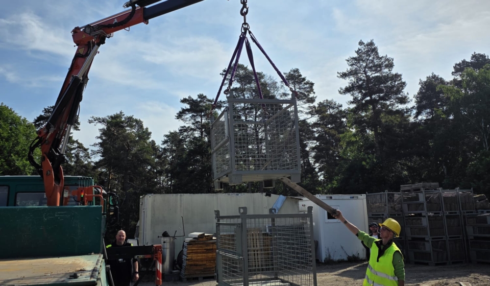 Ein Teilnehmer platziert eine Gitterbox präzise über zwei anderen, während ein Einweiser den Prozess mit einer Holzstange unterstützt.